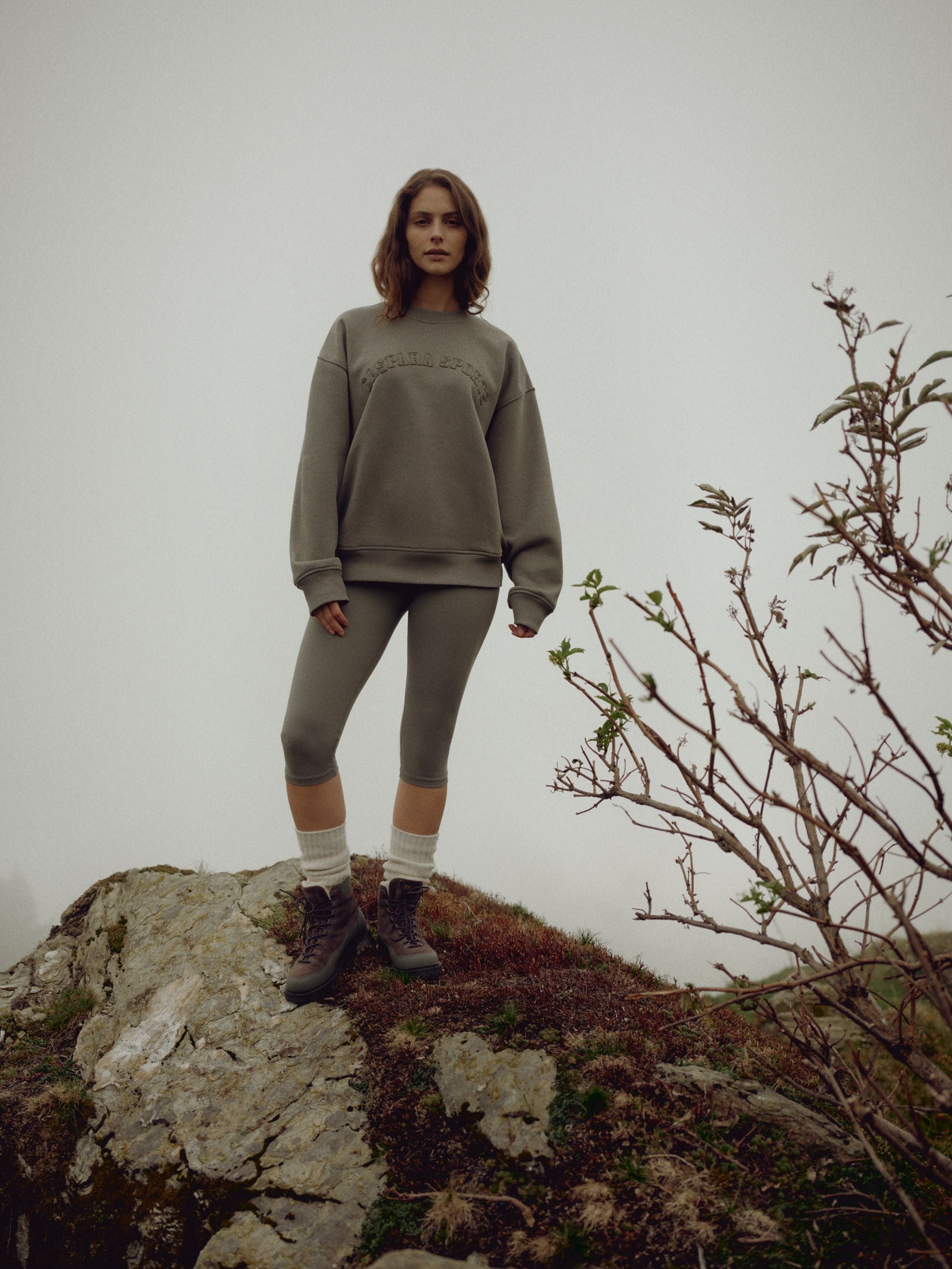 Woman wearing a khaki CASPARA CS crewneck sweatshirt standing on a rocky hill on a foggy day.